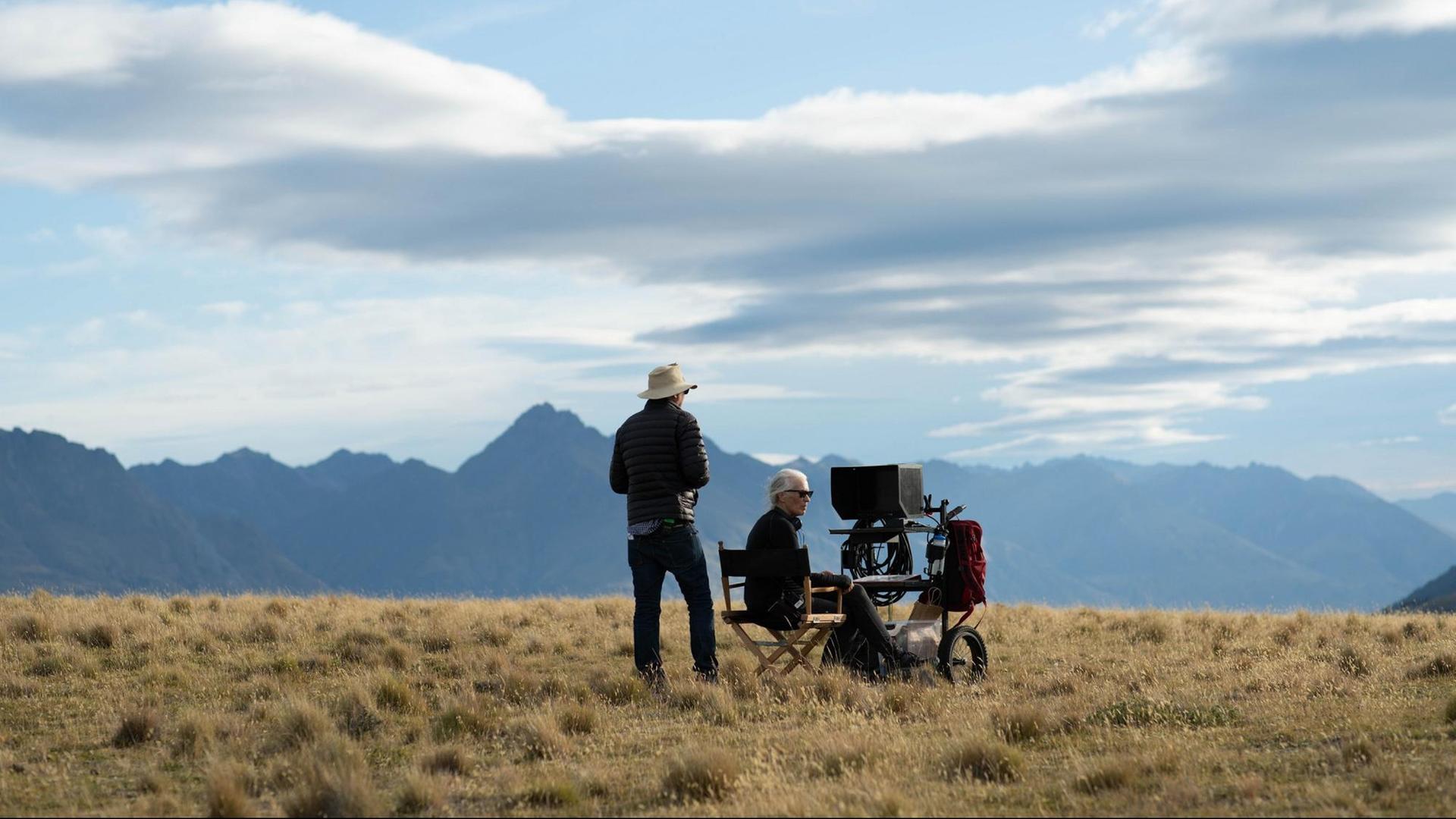 Auf einem Feld sitzt die Regisseurin Jane Campion auf einem Klappstuhl, vor ihr ein Monitor, im Hintergrund die Berge. Neben ihr steht ein Mann mit Cowboyhut Auf einem Feld sitzt die Regisseurin Jane Campion auf einem Klappstuhl, vor ihr ein Monitor, im Hintergrund die Berge. Neben ihr steht ein Mann mit Cowboyhut