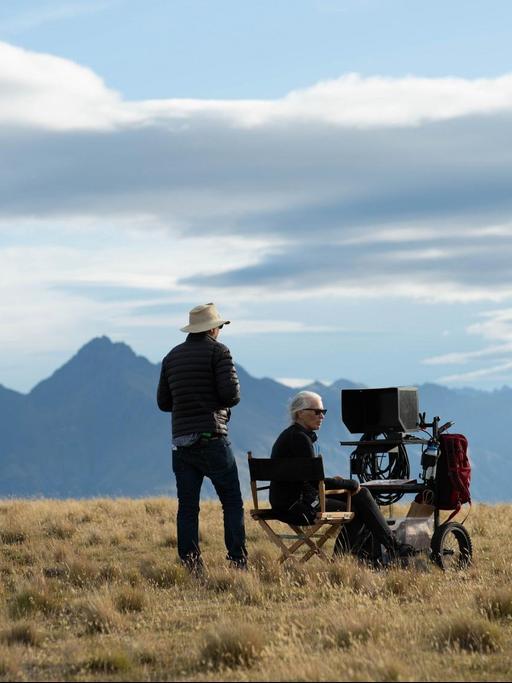 Auf einem Feld sitzt die Regisseurin Jane Campion auf einem Klappstuhl, vor ihr ein Monitor, im Hintergrund die Berge. Neben ihr steht ein Mann mit Cowboyhut