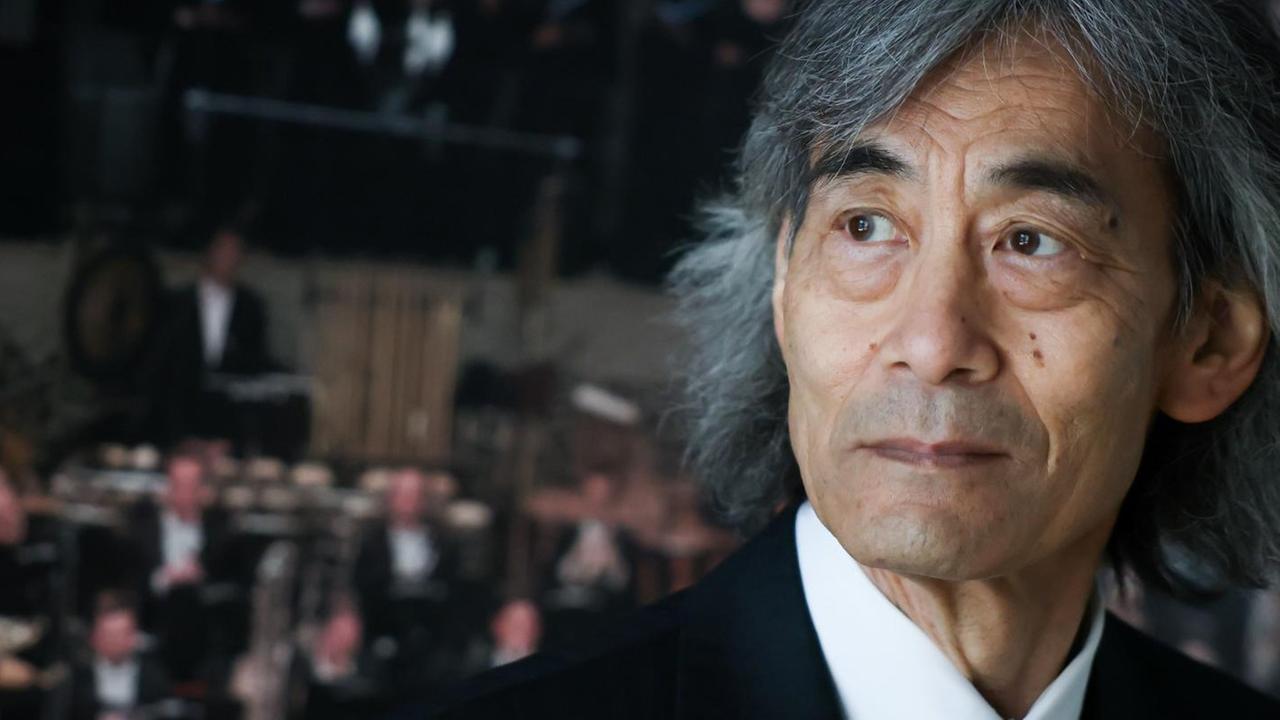 Kent Nagano steht vor einem Poster, das Musiker in den Rängen der Elbphilharmonie zeigt. Kent Nagano steht vor einem Poster, das Musiker in den Rängen der Elbphilharmonie zeigt.