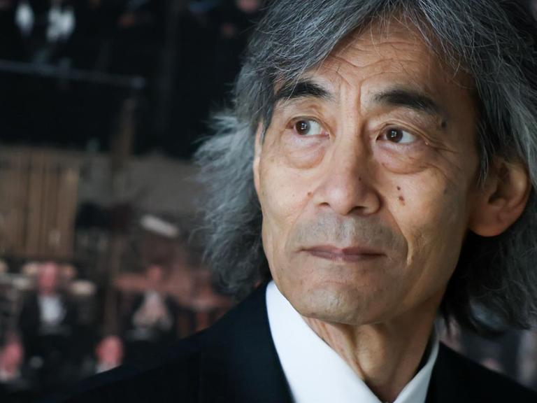 Kent Nagano steht vor einem Poster, das Musiker in den Rängen der Elbphilharmonie zeigt.
