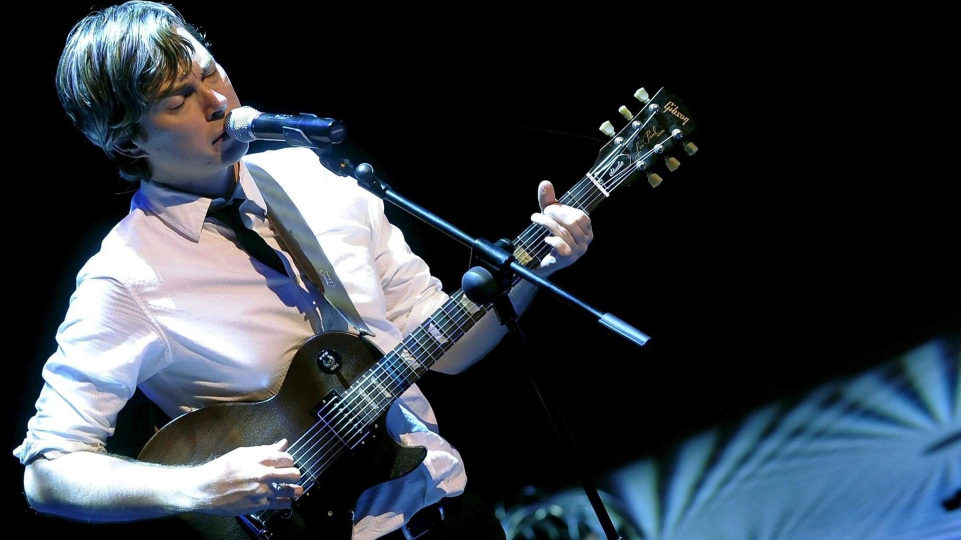 Auf einem schräg fotografierten Bild ist ein Mann im weißen Hemd zu sehen, der Gitarre spielt und in ein Mikrofon singt