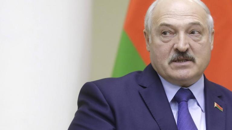 Lukaschenko sitzt in einem Stuhl und debattiert vor einem Hintergrund mit mehreren Flaggen.