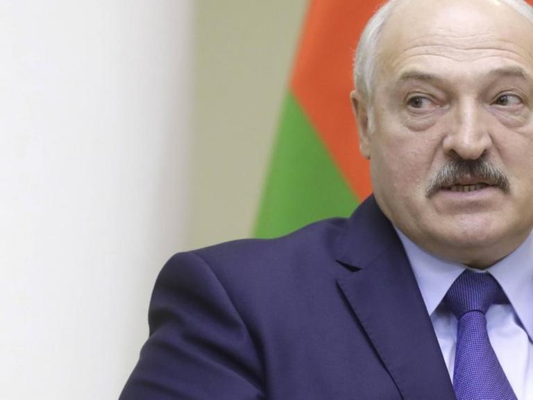 Lukaschenko sitzt in einem Stuhl und debattiert vor einem Hintergrund mit mehreren Flaggen.