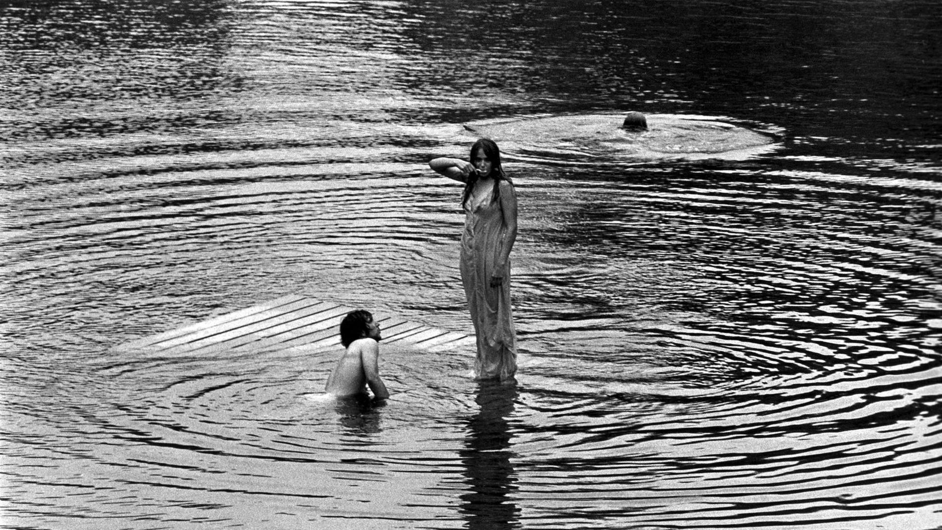 Schwarz-Weiß-Fotografie einer Frau, die im nassen Kleid auf dem Wasser zu stehen scheint, neben ihr schwimmen zwei Männer im See. Schwarz-Weiß-Fotografie einer Frau, die im nassen Kleid auf dem Wasser zu stehen scheint, neben ihr schwimmen zwei Männer im See.