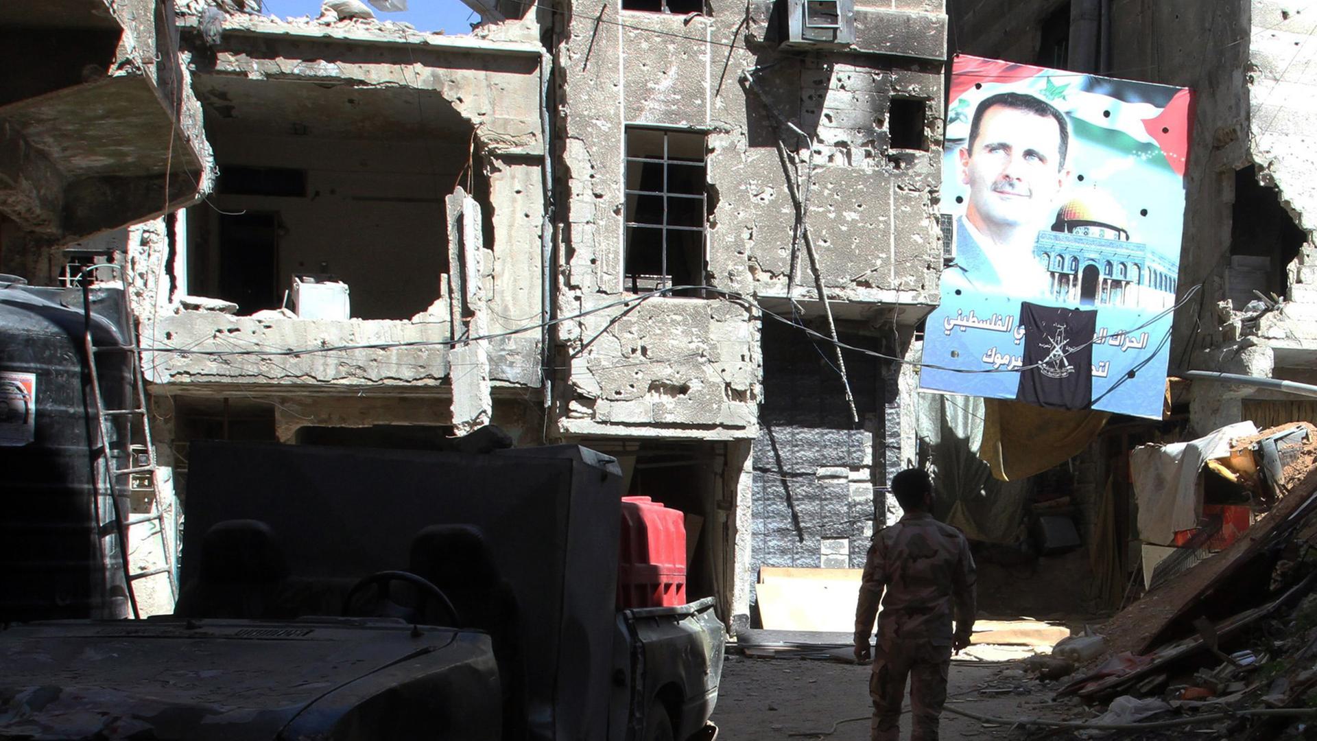 Zwischen beschädigten Häusern geht ein Mann durch die Straßen des Flüchtlingslagers, vor ihm ein großes Plakat mit einem Porträt des syrischen Präsidenten Baschar al-Assad. Zwischen beschädigten Häusern geht ein Mann durch die Straßen des Flüchtlingslagers, vor ihm ein großes Plakat mit einem Porträt des syrischen Präsidenten Baschar al-Assad.