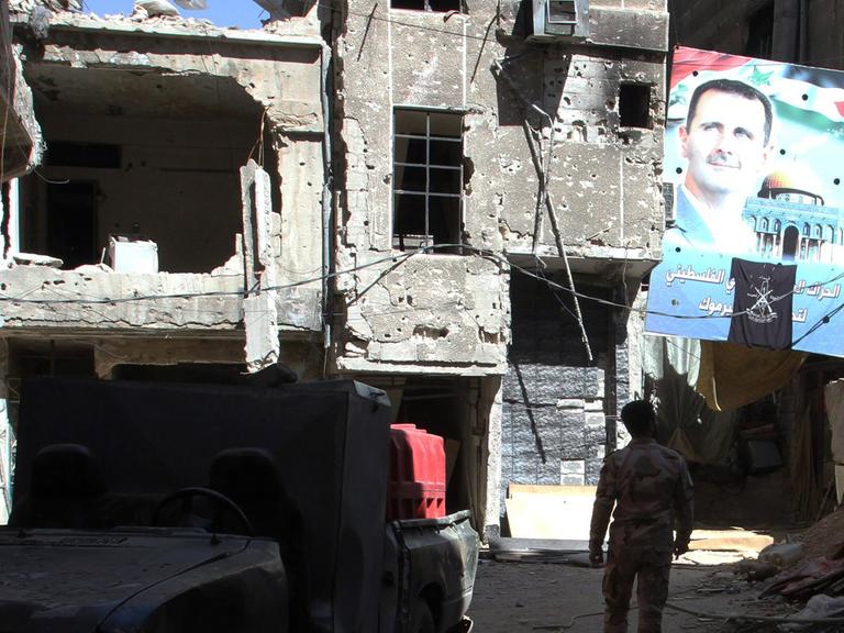 Zwischen beschädigten Häusern geht ein Mann durch die Straßen des Flüchtlingslagers, vor ihm ein großes Plakat mit einem Porträt des syrischen Präsidenten Baschar al-Assad.
