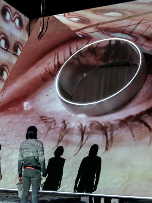 Das Foto aus dem Theaterstück "Evolution" zeigt ein überdimensioniertes Auge vor dem zwei Menschen, mit dem Rücken zum Publikum, stehen