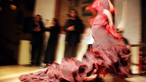 Eine in rot gekleidete Flamencotänzerin. Im Hintergrund schauen ihr drei Personen beim Tanzen zu.