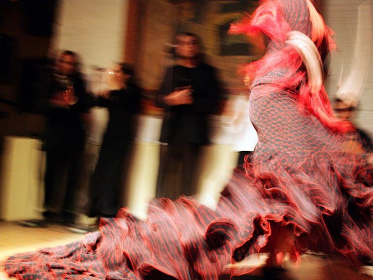 Eine in rot gekleidete Flamencotänzerin. Im Hintergrund schauen ihr drei Personen beim Tanzen zu.