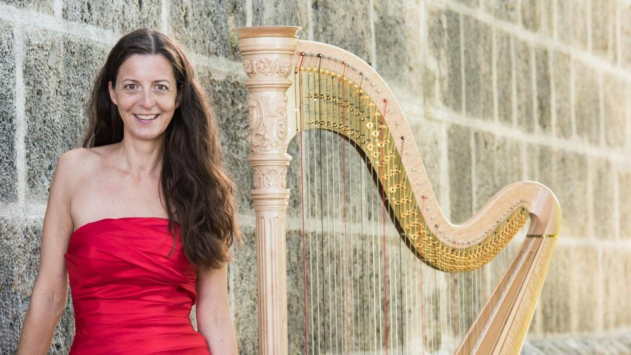 Eine Frau steht in rotem Kleid neben ihrer Harfe, beide vor einer massiven, steinernen Mauer. Eine Frau steht in rotem Kleid neben ihrer Harfe, beide vor einer massiven, steinernen Mauer.