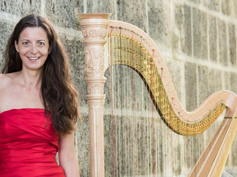 Die Musikerin steht in rotem Kleid neben ihrer Harfe, beide vor einer massiven, steinernen Mauer.