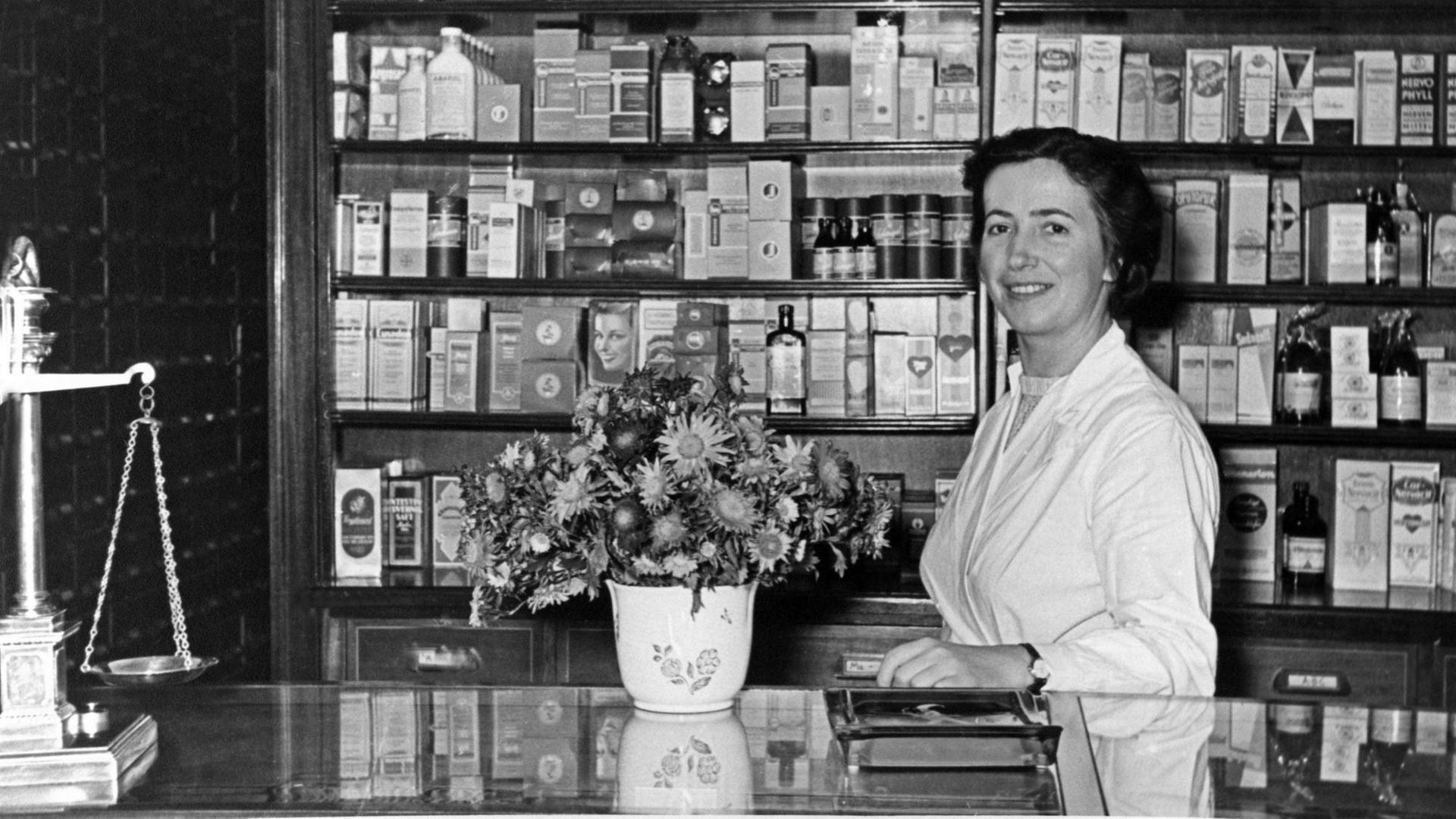 Eine historische schwarz-weiß-Aufnahme einer typischen deutschen Apotheke aus den 50er Jahren zeigt eine Apothekeri in weißem Kittel hinter einem Tresen auf dem ein Blumenstrauß und eine Waage im Anschnitt zu sehen sind.