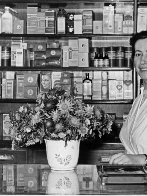 Eine historische schwarz-weiß-Aufnahme einer typischen deutschen Apotheke aus den 50er Jahren zeigt eine Apothekeri in weißem Kittel hinter einem Tresen auf dem ein Blumenstrauß und eine Waage im Anschnitt zu sehen sind.
