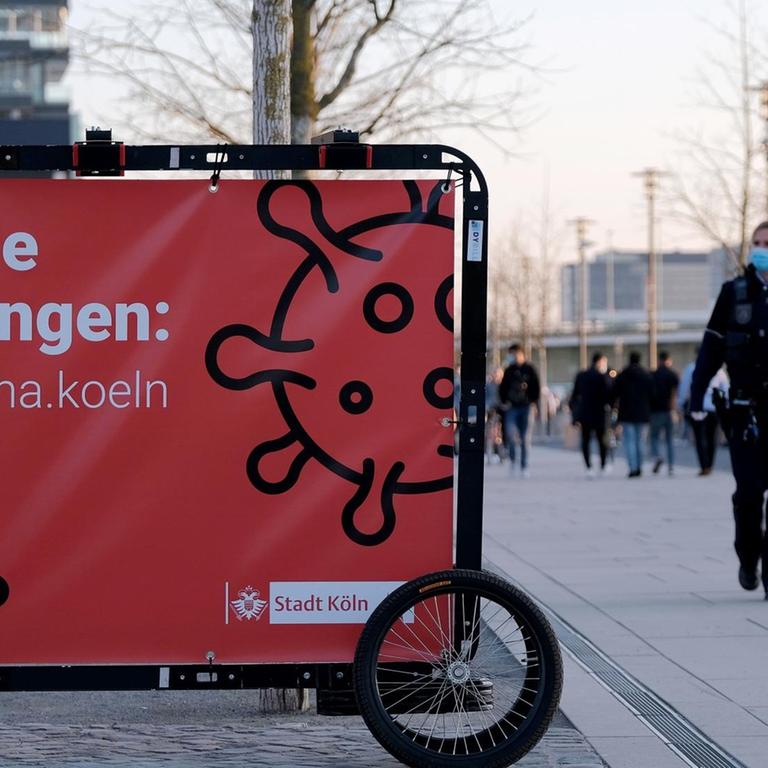 Ein fahrbares Transparent mit der Aufschrift "Aktuelle Regelungen" steht auf dem Rheinboulevard in Köln. Daneben gehen zwei Polizisten mit Masken. 