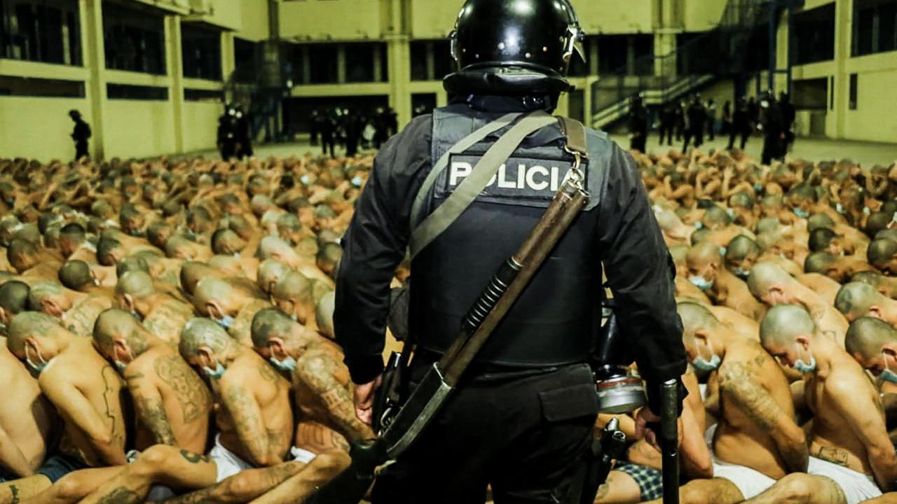 Halbnackte Gefangene sitzen in engen Reihen im Innenhof eines Gefängnisses, im Vordergrund von einem bewaffneten Polizisten bewacht. Halbnackte Gefangene sitzen in engen Reihen im Innenhof eines Gefängnisses, im Vordergrund von einem bewaffneten Polizisten bewacht.