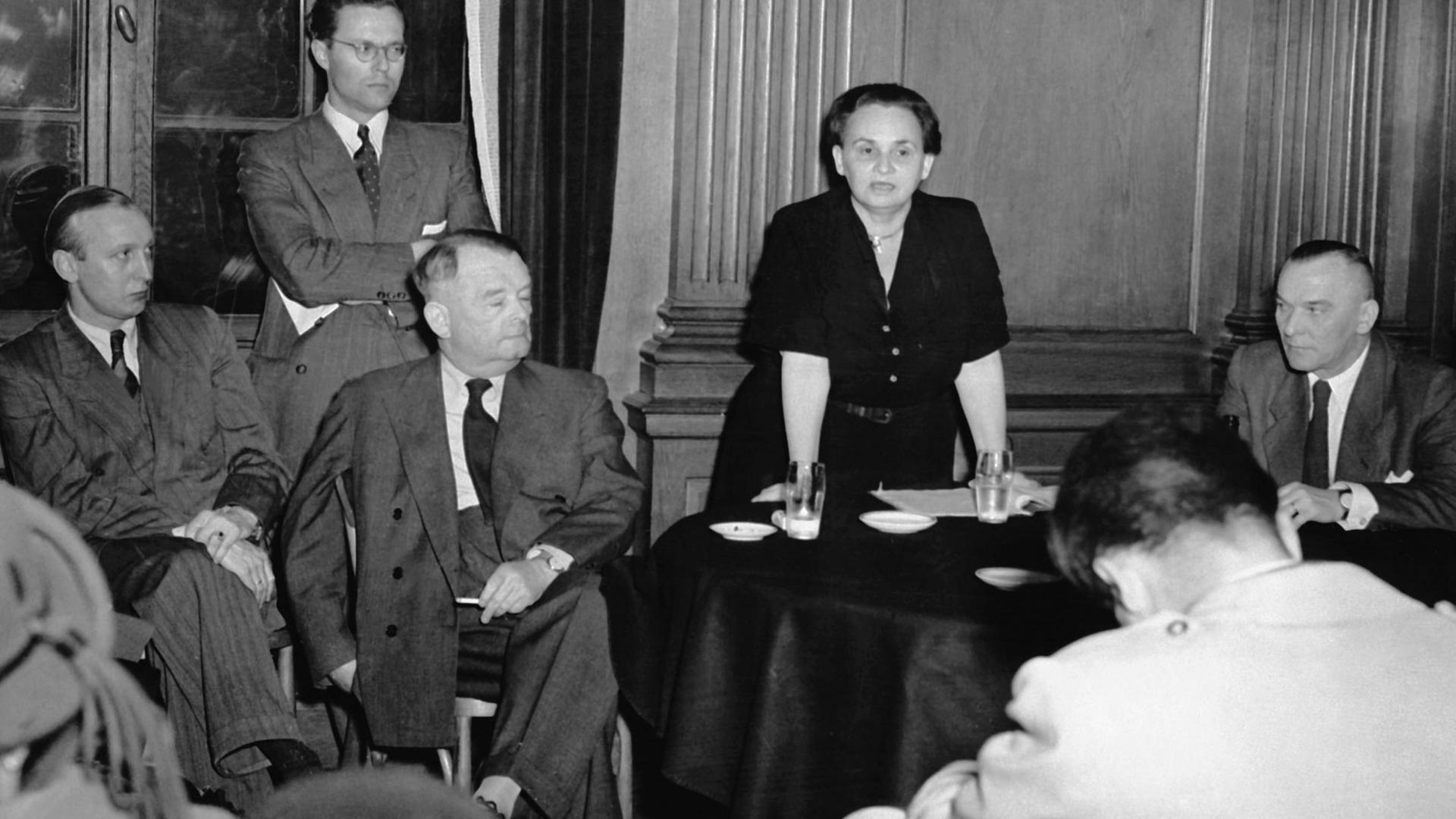Ein schwarzweißes Foto zeigt die ehemalige deutsche Kommunistin Ruth Fischer während eines Vortrags im Mai 1951 in Frankfurt am Main, auf einen Tisch gestützt, umgeben von fünf  Männern in Anzügen