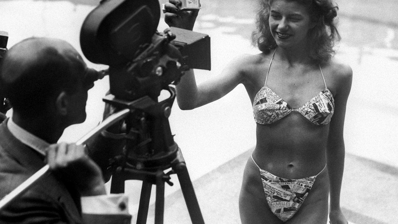 Ein Schwarz-Weiß-Bild zeigt einen Fotografen, der durch eine Kamera auf das Model Micheline Bernardini blickt, die am 5. Juli 1946 in einem Schwimmbad den ersten Bikini vorführt Ein Schwarz-Weiß-Bild zeigt einen Fotografen, der durch eine Kamera auf das Model Micheline Bernardini blickt, die am 5. Juli 1946 in einem Schwimmbad den ersten Bikini vorführt
