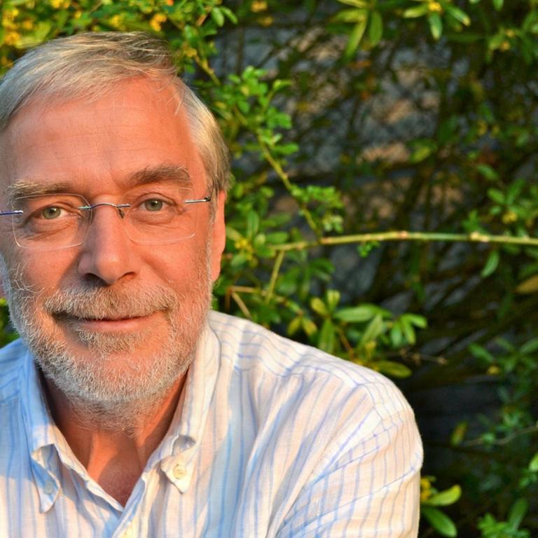Der Neurobiologe Gerald Hüther, fotografiert in einem hell gestreifen Hemd vor einer grünen Hecke