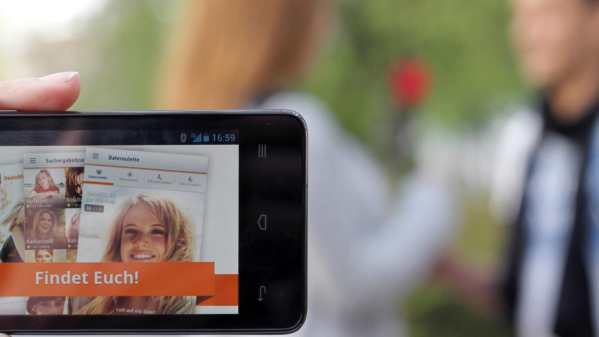 Ein Handy, in dessen Hintergrund sich zwei Menschen eine Rose reichen Ein Handy, in dessen Hintergrund sich zwei Menschen eine Rose reichen