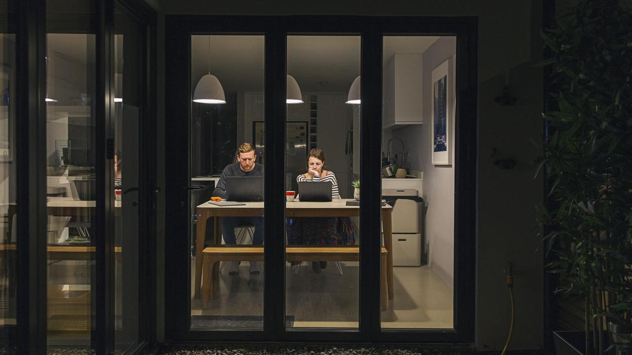Blick bei Nacht in eine beleuchtete Küche. Ein Paar sitzt mit Laptops am Küchentisch. Blick bei Nacht in eine beleuchtete Küche. Ein Paar sitzt mit Laptops am Küchentisch.