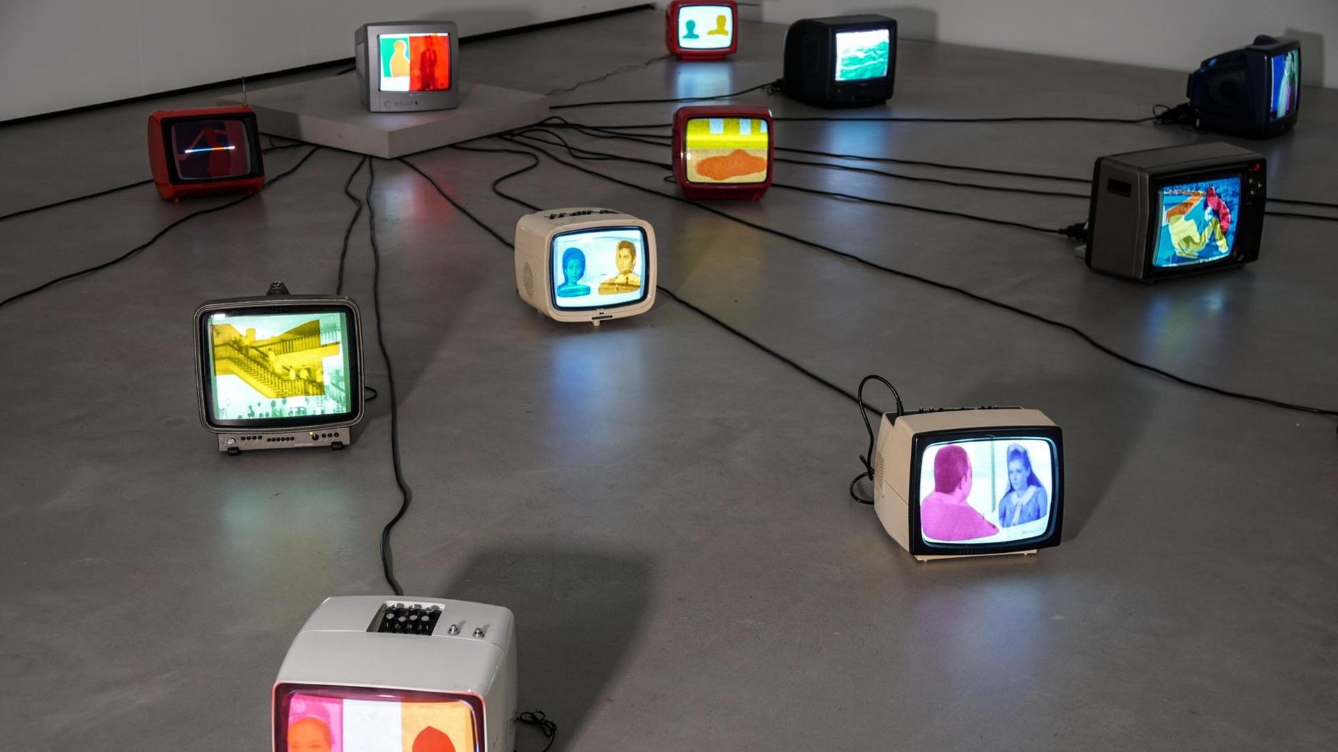 Auf dem Boden stehen verschiedene alte Fernseher, die mit Kabeln zu einem zentralen Fernseher verbunden sind. Auf die Monitore sind gefärbte Folien geklebt. Auf dem Boden stehen verschiedene alte Fernseher, die mit Kabeln zu einem zentralen Fernseher verbunden sind. Auf die Monitore sind gefärbte Folien geklebt.