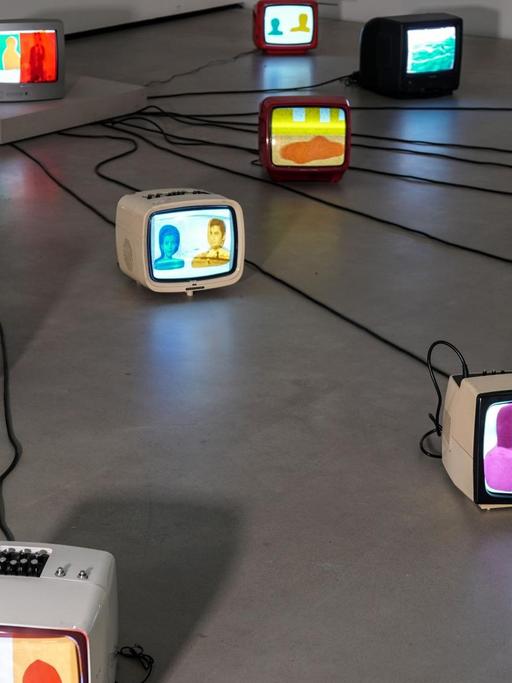 Auf dem Boden stehen verschiedene alte Fernseher, die mit Kabeln zu einem zentralen Fernseher verbunden sind. Auf die Monitore sind gefärbte Folien geklebt.