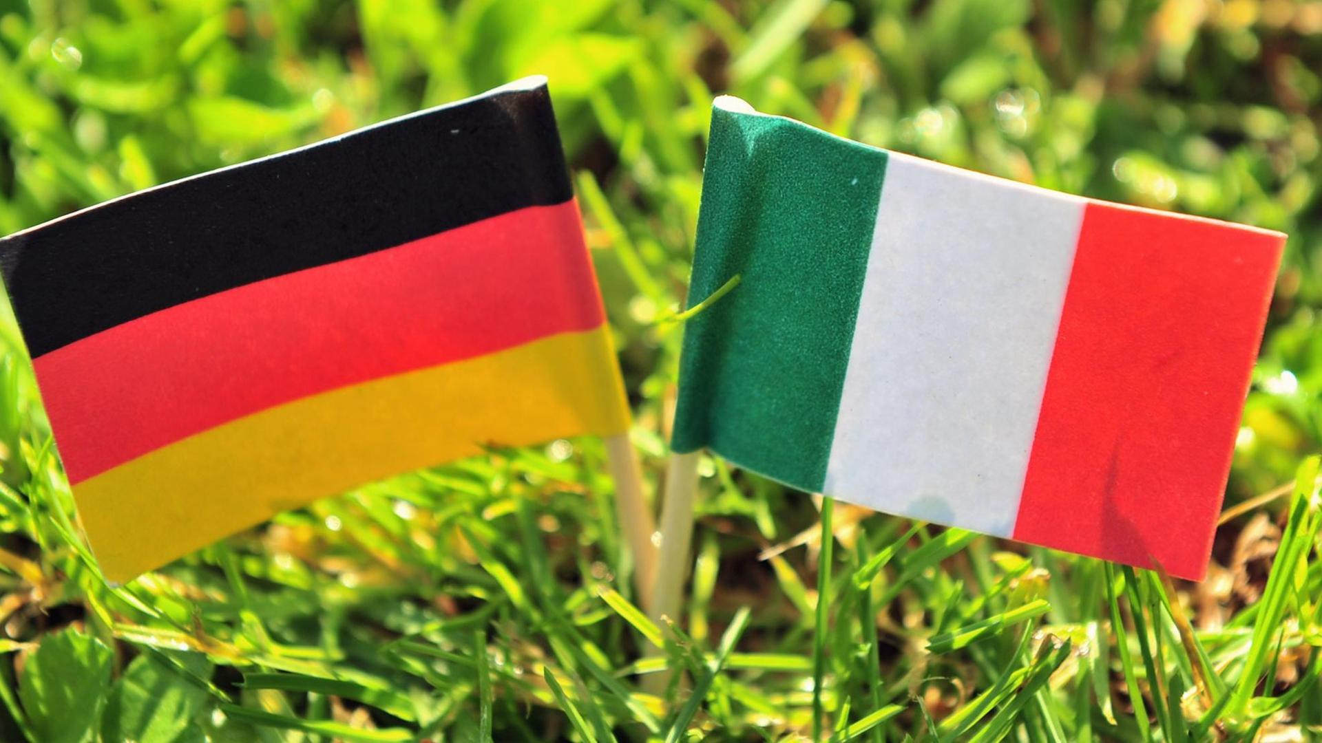 Die deutsche und die italienische Flagge stecken im Rasen eines Gartens.