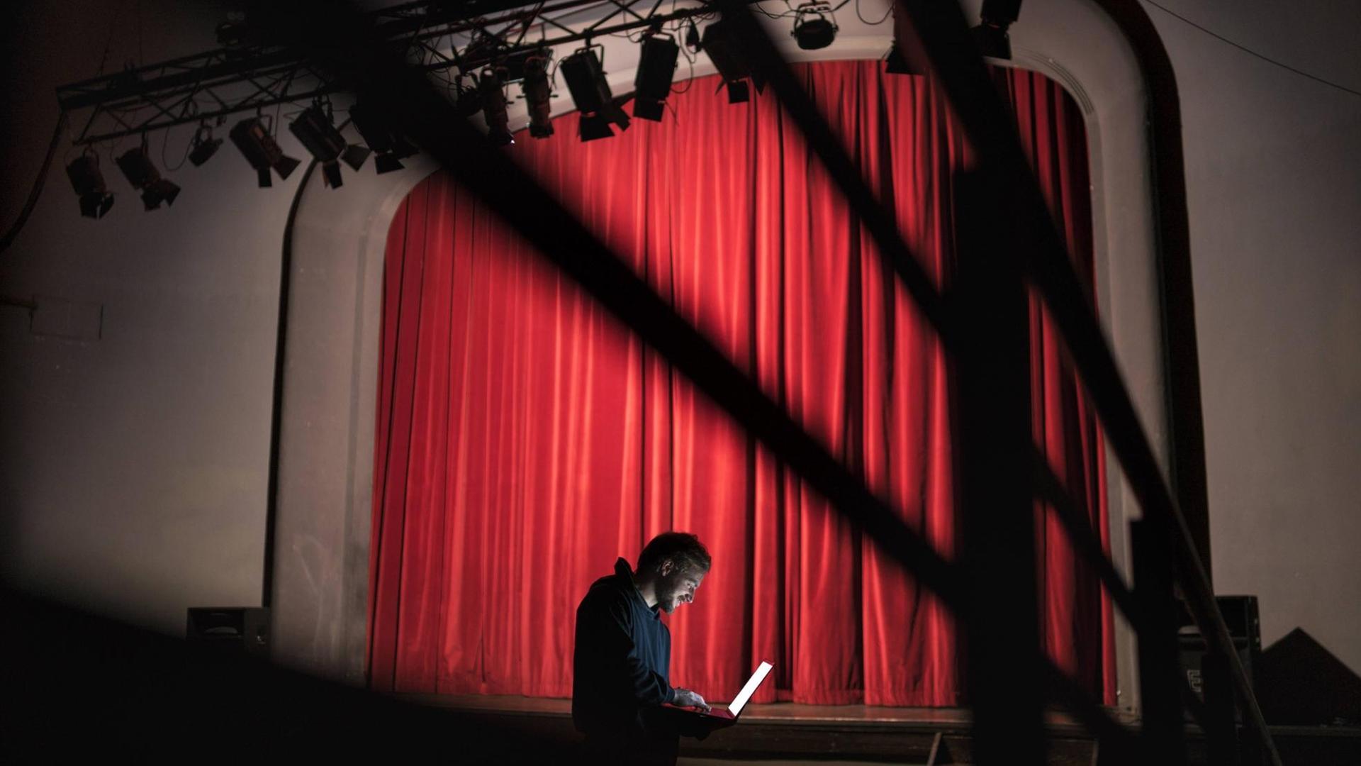 Ein Regisseur schaut im Zuschauerraum eines Theaters auf einen Laptop. Im Hintergrund: die Bühne mit geschlossenem Vorhang. Ein Regisseur schaut im Zuschauerraum eines Theaters auf einen Laptop. Im Hintergrund: die Bühne mit geschlossenem Vorhang.