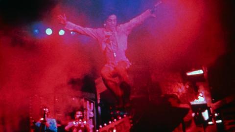 Prince ist auf der Bühne, eingetaucht in rotem und blauen Licht, in Nebelschwaden zu sehen. Er hat keinen Boden unter den Füßen.