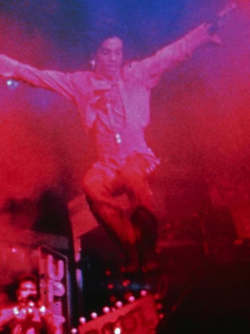 Prince ist auf der Bühne, eingetaucht in rotem und blauen Licht, in Nebelschwaden zu sehen. Er hat keinen Boden unter den Füßen.