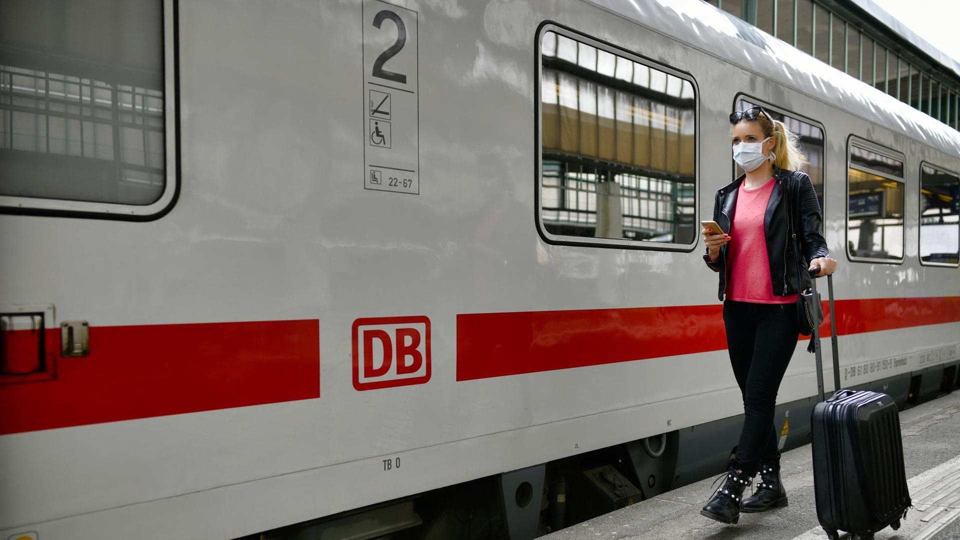 Eine Frau mit einer Maske steht am Bahn-Hof neben einem Zug. Sie hat ein Handy in der Hand. Eine Frau mit einer Maske steht am Bahn-Hof neben einem Zug. Sie hat ein Handy in der Hand.