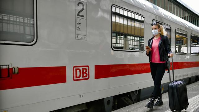 Eine Frau mit einer Maske steht am Bahn-Hof neben einem Zug. Sie hat ein Handy in der Hand.