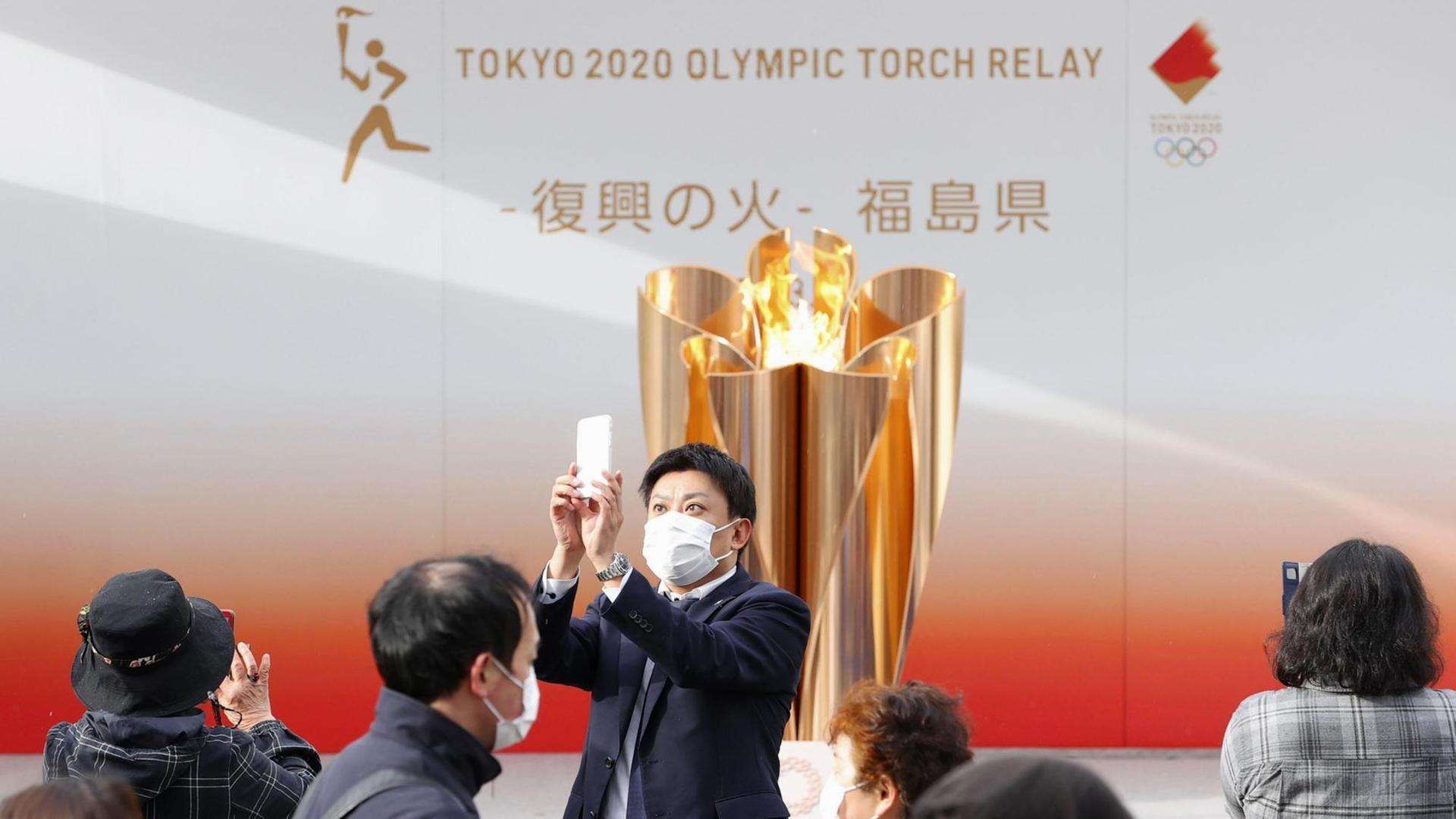 24.03.2020, Japan, Fukushima: Ein Besucher mit Mundschutz macht ein Selfie mit der olympischen Flamme in Fukushima. Die Flamme tourt durch die von der Erdbeben-Tsunami-Katastrophe im März 2011 schwer getroffenen Gebiete im Nordosten Japans, bevor der Fackellauf in der Präfektur Fukushima am 26. März beginnt. Foto: kyodo/dpa +++ dpa-Bildfunk +++ 24.03.2020, Japan, Fukushima: Ein Besucher mit Mundschutz macht ein Selfie mit der olympischen Flamme in Fukushima. Die Flamme tourt durch die von der Erdbeben-Tsunami-Katastrophe im März 2011 schwer getroffenen Gebiete im Nordosten Japans, bevor der Fackellauf in der Präfektur Fukushima am 26. März beginnt. Foto: kyodo/dpa +++ dpa-Bildfunk +++