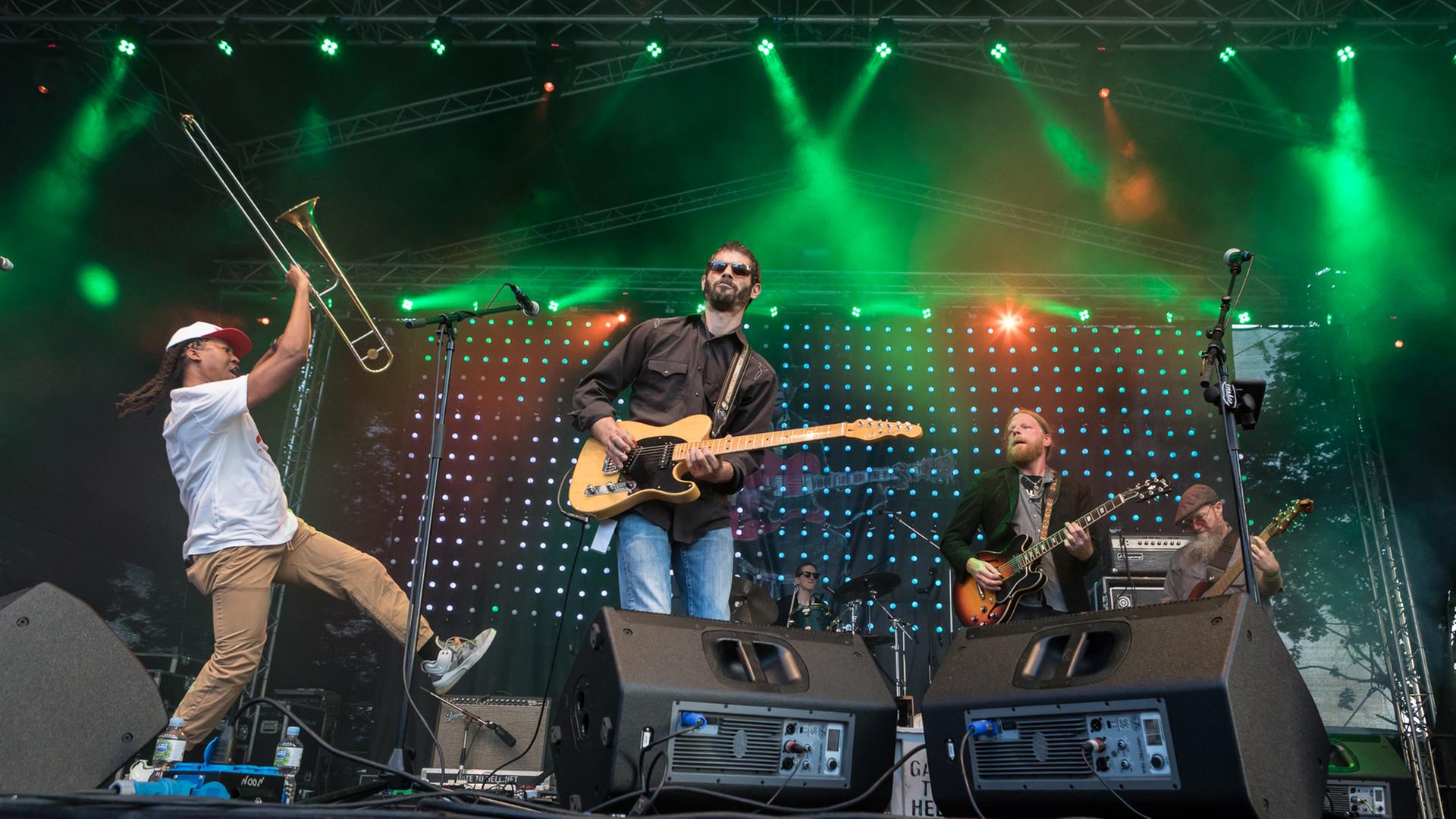 Zwei Gitarristen, rechts ein Bassist, hinten ein Schlagzeuger auf einer Bühne, vorne links springt ein Mann mit einer Posaune in der Hand in die Luft. Zwei Gitarristen, rechts ein Bassist, hinten ein Schlagzeuger auf einer Bühne, vorne links springt ein Mann mit einer Posaune in der Hand in die Luft.