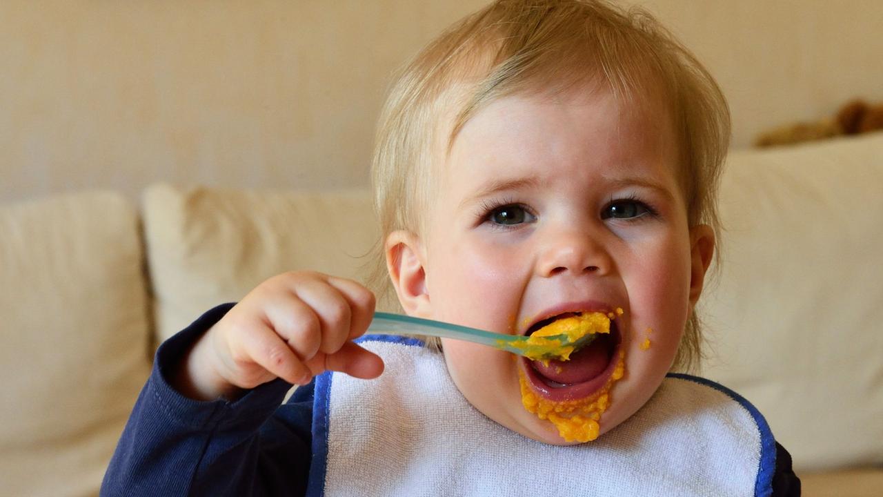 Ein kleines Mädchen löffelt ohne Hilfe allein und mit großem Appetit seinen Möhrenbre aus einer Plastikschüsseli.