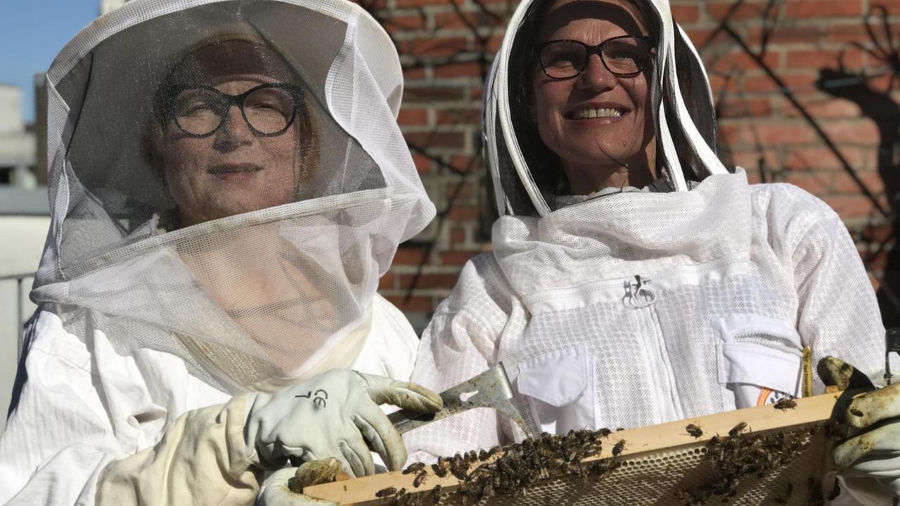 Das Bild zeigt die beiden Imkerinnen Iris Pinkepank und Stephanie Breil vom Projekt "HonigConnection" im Imkerschutzanzug mit einem Wabenrähmchen voller Bienen in den Händen. Das Bild zeigt die beiden Imkerinnen Iris Pinkepank und Stephanie Breil vom Projekt "HonigConnection" im Imkerschutzanzug mit einem Wabenrähmchen voller Bienen in den Händen.