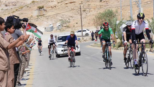 Kinder mit jordanischen Fahnen stehen am Rande einer Radstrecke, Rennradfahrer fahren vorbei.