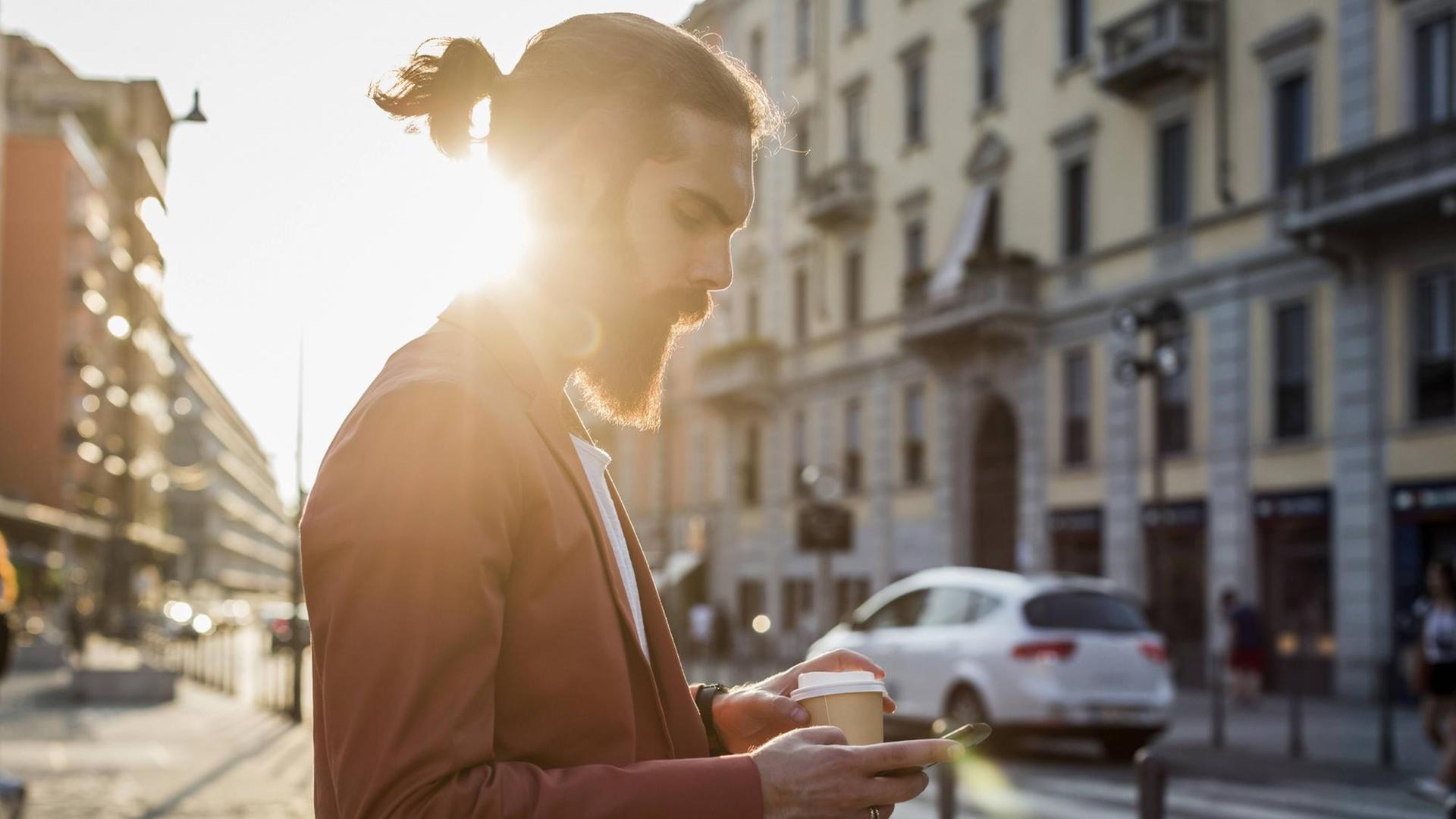 Ein junger Mann mit Vollbart geht mir einem Kaffee in der Hand über eine Straße und Blick auf sein Handy. Ein junger Mann mit Vollbart geht mir einem Kaffee in der Hand über eine Straße und Blick auf sein Handy.