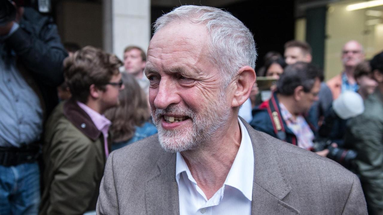 Corbyn im grauen Anzug auf einer Straße, hinter einer Absperrung stehen Fotografen. Corbyn im grauen Anzug auf einer Straße, hinter einer Absperrung stehen Fotografen.