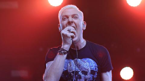 Ein Mann mit kurzen blonden Haaren steht auf einer Bühne und schreit in ein Mikrofon.