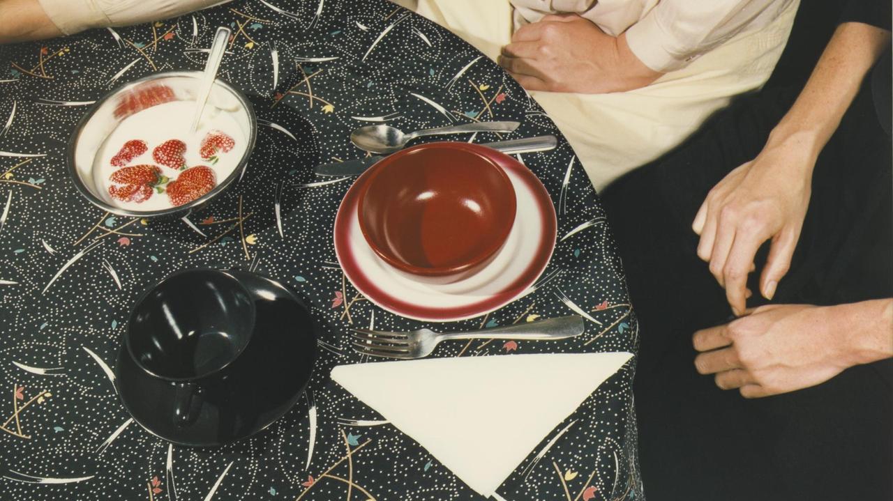 Ein ausgesucht gedeckter Tisch mit rot-weißem und schwarzem Geschirr neben einer Schale mit Erdbeeren in weißer Flüssigkeit auf einer gemusterten Tischdecke Ein ausgesucht gedeckter Tisch mit rot-weißem und schwarzem Geschirr neben einer Schale mit Erdbeeren in weißer Flüssigkeit auf einer gemusterten Tischdecke