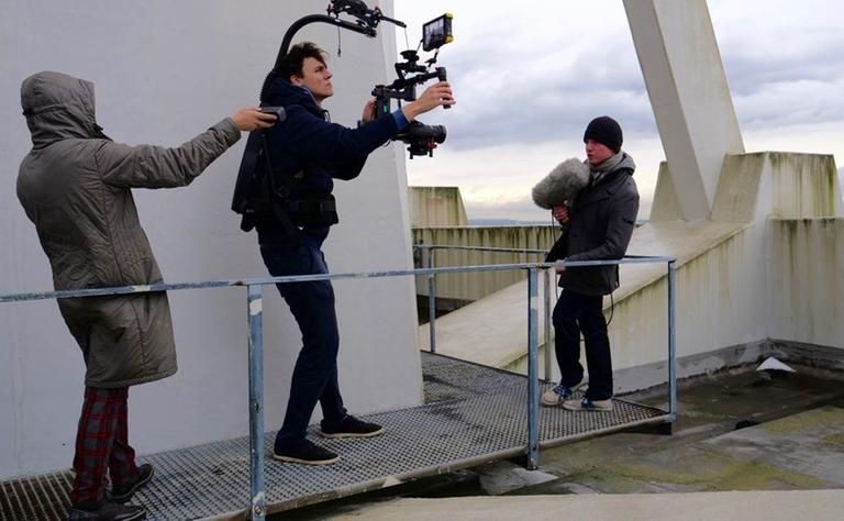 Martin Zingsheim auf dem Dach des Deutschlandfunk-Hauses. vor ihm ein Kameramann, der ihn filmt und dahinter eine Person, die bei dem Videodreh assistiert.