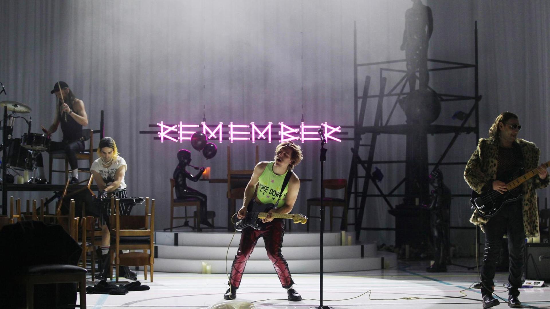 Szenenfoto aus "In my Room": Eine Band spielt auf einer Bühne. Im Vordergrund Jonas Dassler in einem neongrünen bauchfreien Muskel-Shirt mit dem Aufdruck "Upside Down". Er spielt Gitarre.