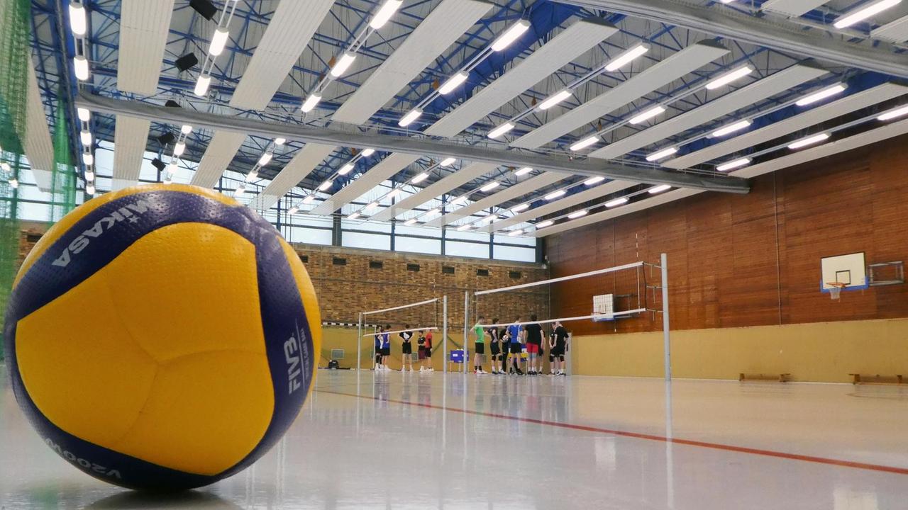 Sporthalle mit Spielern, die sich am Spielfeld bespechen. Im Vordergrund ist ein gelb-blauer Volleyball zu sehen. Sporthalle mit Spielern, die sich am Spielfeld bespechen. Im Vordergrund ist ein gelb-blauer Volleyball zu sehen.