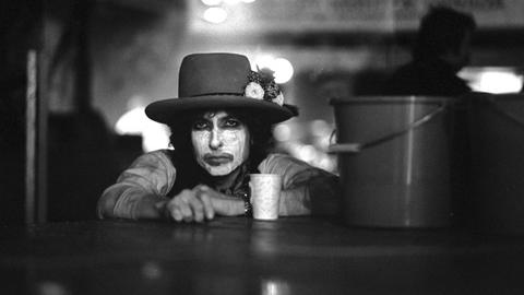 Der Musiker Bob Dylan, die Hände auf einer Tischplatte, mit weiß geschminktem Gesicht und dunklem Hut sowie mit einem Kaffeebecher vor und einem Putzeimer neben sich.