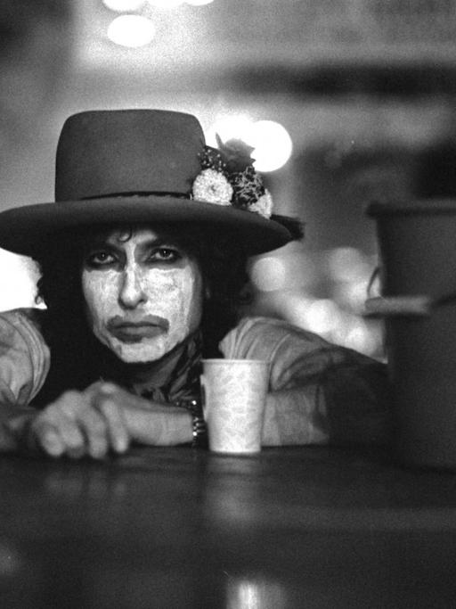 Der Musiker Bob Dylan, die Hände auf einer Tischplatte, mit weiß geschminktem Gesicht und dunklem Hut sowie mit einem Kaffeebecher vor und einem Putzeimer neben sich.