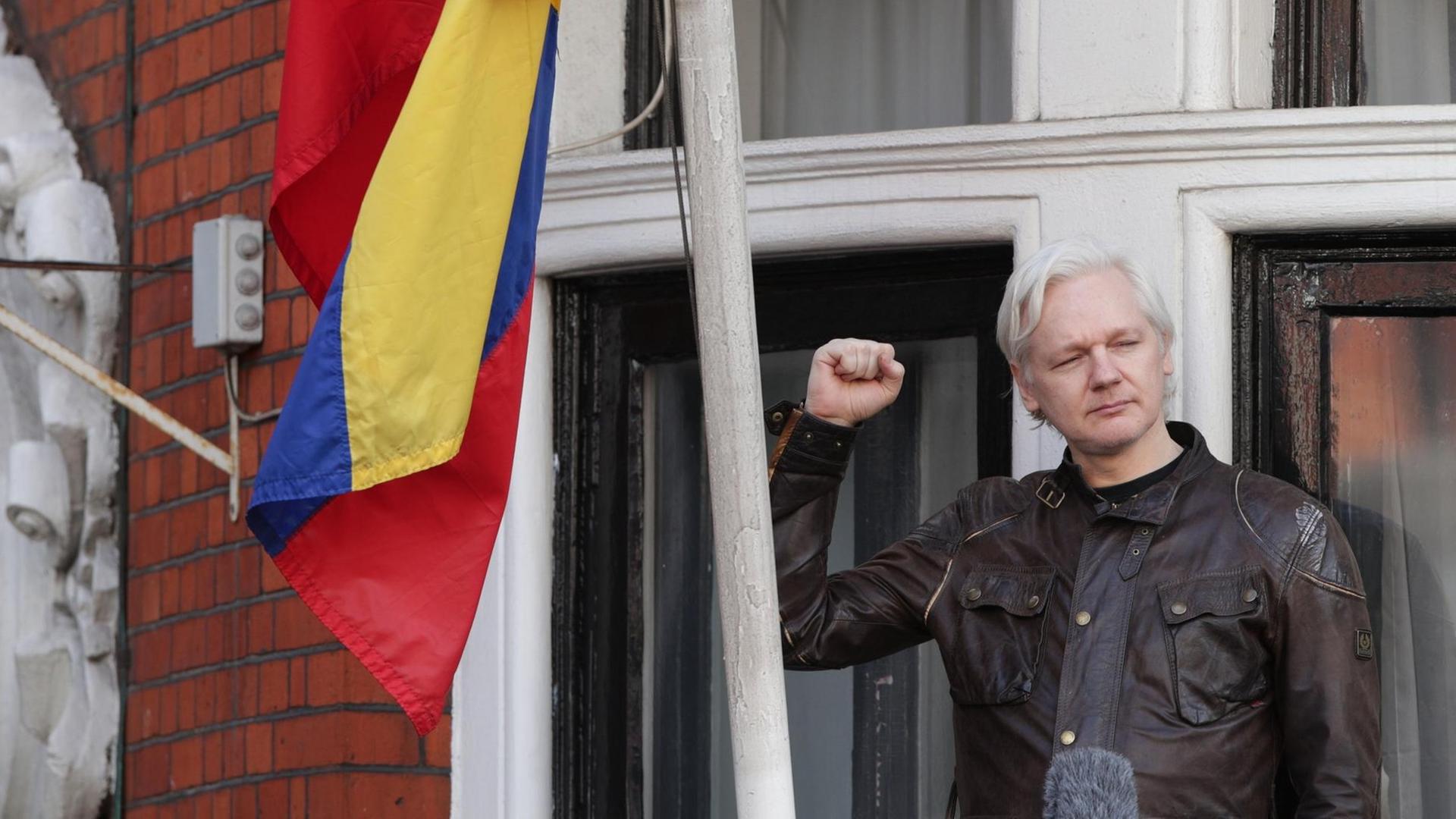 Assange trägt eine schwarze Jacke und hebt kämpferisch die Faust. Neben ihm die Fahne Ecuadors.