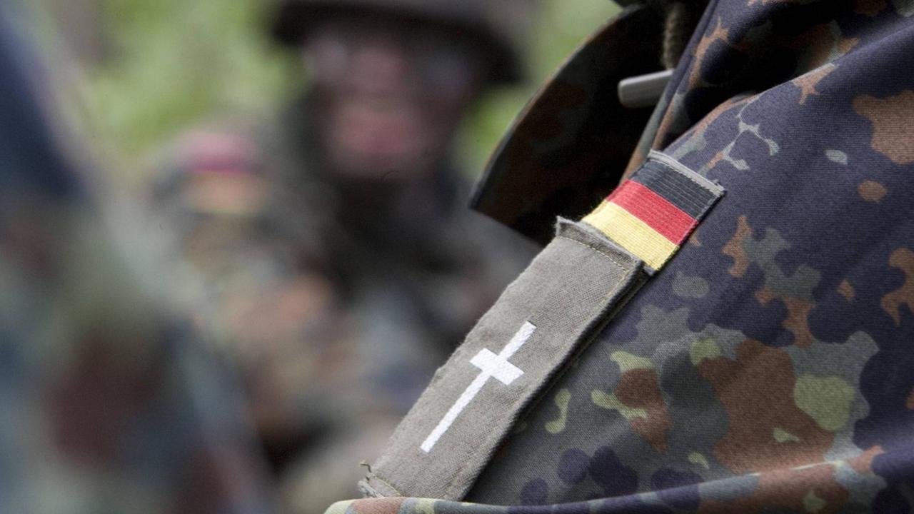 Schulterklappe mit Kreuz eines evangelischen Militärpfarrers unter einem Aufneher einer Deutschlandfahne auf der Schulter einer Camouflage Jacke Schulterklappe mit Kreuz eines evangelischen Militärpfarrers unter einem Aufneher einer Deutschlandfahne auf der Schulter einer Camouflage Jacke
