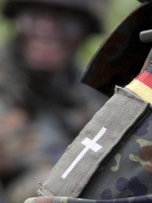 Schulterklappe mit Kreuz eines evangelischen Militärpfarrers unter einem Aufneher einer Deutschlandfahne auf der Schulter einer Camouflage Jacke