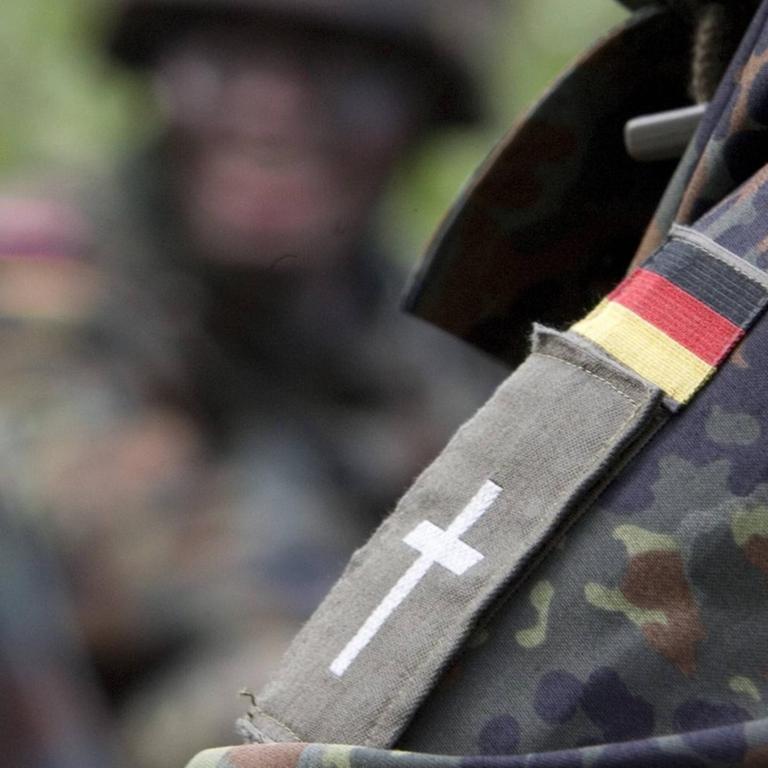Schulterklappe mit Kreuz eines evangelischen Militärpfarrers unter einem Aufneher einer Deutschlandfahne auf der Schulter einer Camouflage Jacke